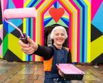 woman stood infront of a brightly painted wall, holding a paint roller covered in light pink paint