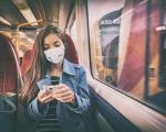 young woman sat on a train wearing a protective mask while looking at her phone