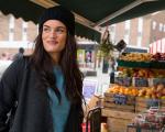 woman with long dark hair in a beanie hat smiling at a fruit market
