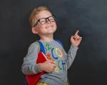 young blonde boy wearing a red backpack while smiling and pointing in the air.