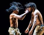 Two long-haired men dancing shirtless against a black background