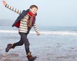 boy in a long sleeved red and white top and a red scarf running along the beach with his arms spread out