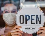 person in a white face mask holding a shop sign that says 'open'
