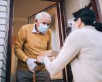 lady wearing a facemask passing a boxed meal to an elderly man at his front door