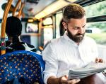 bearded man in a white shirt sat on a bus reading a newspaper