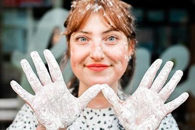 woman with smiling towards the camera with her hands facing forward creating a 'w' shape - her hands and hair have flour on them