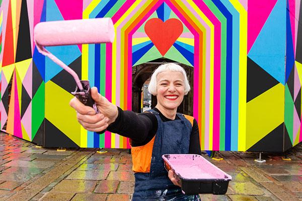 woman standing against a brightly painted wall, holding a paint roller covered in light pink paint