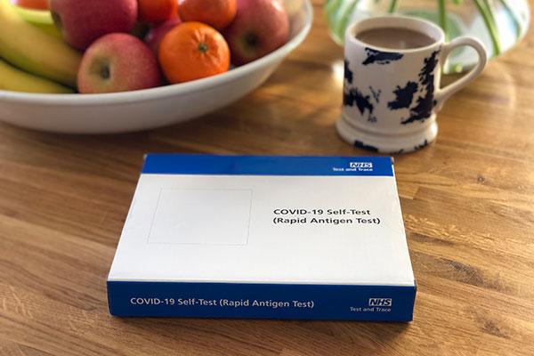 box of Covid-19 rapid tests on a coffee table in front of a bowl of fruit and a hot drink