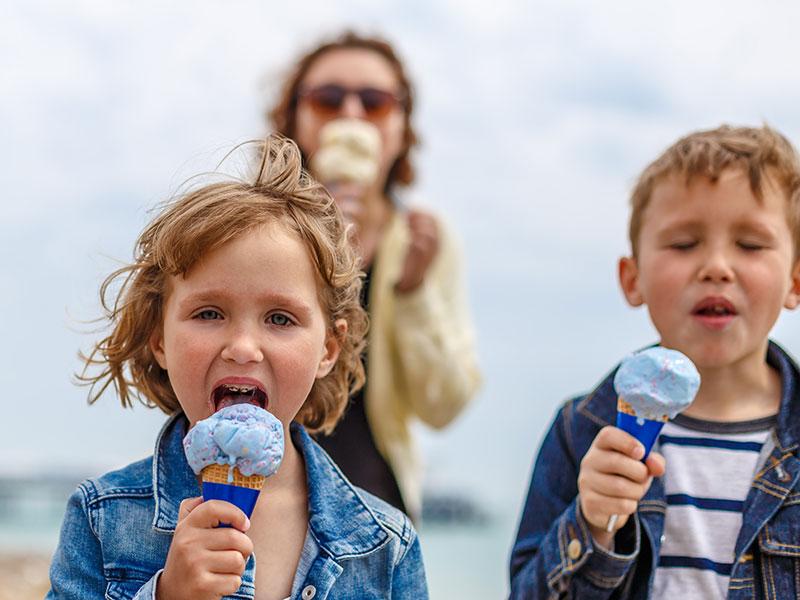 two children eating ice cream on a beach with their mother in the background