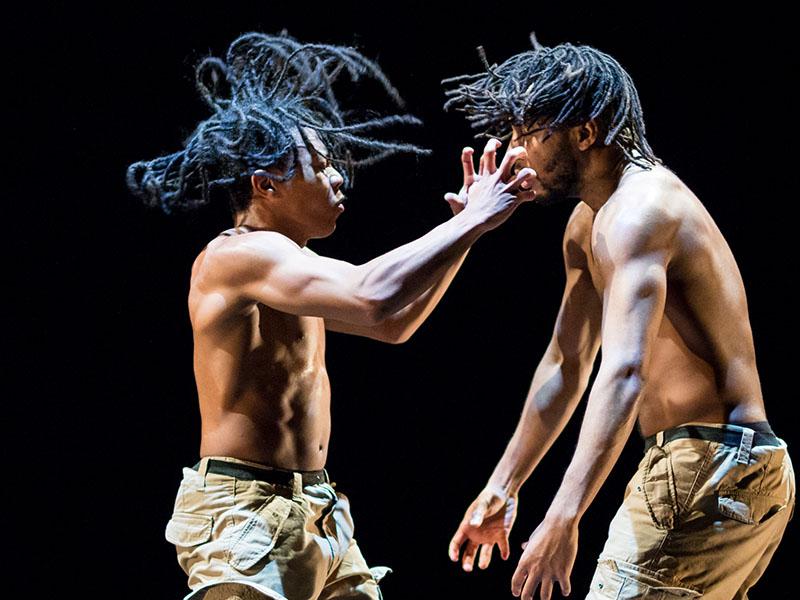 Two long-haired men dancing shirtless against a black background