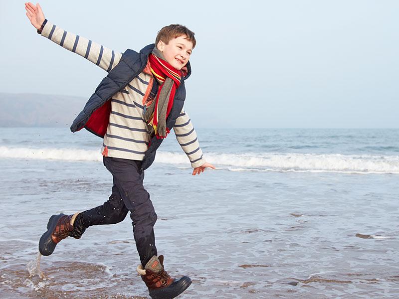 boy in a long sleeved red and white top and a red scarf running along the beach with his arms spread out