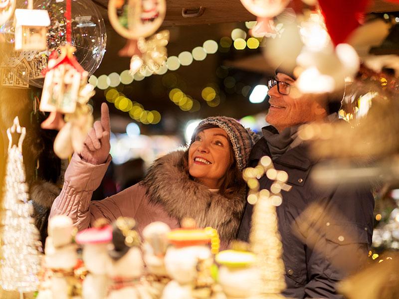 man and woman wearing winter clothes looking at items at a Christmas market stall covered in lights