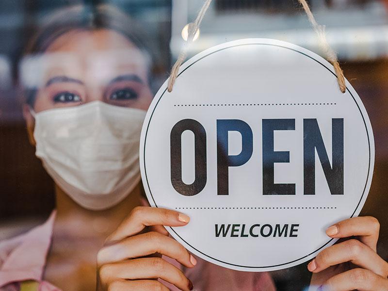 person in a white face mask holding a shop sign that says 'open'