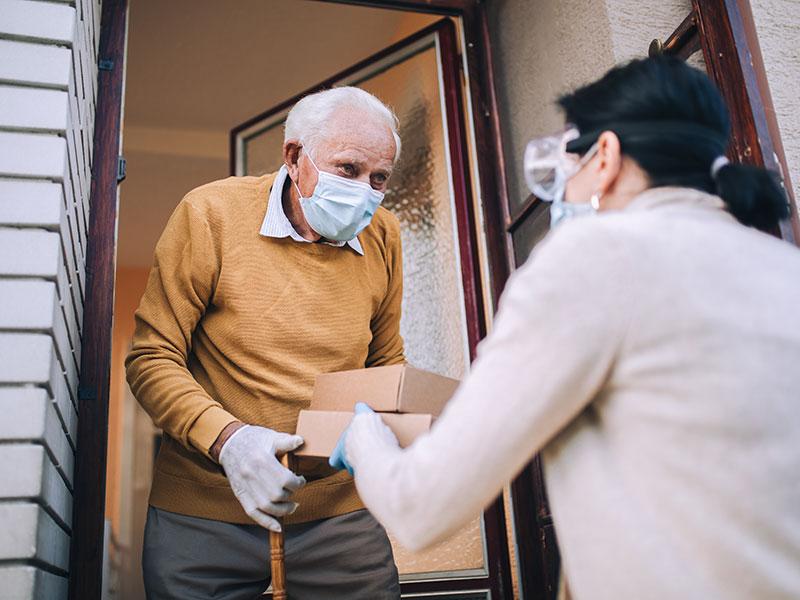 lady wearing a facemask passing a boxed meal to an elderly man at his front door
