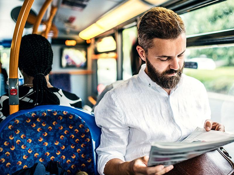 bearded man in a white shirt sat on a bus reading a newspaper