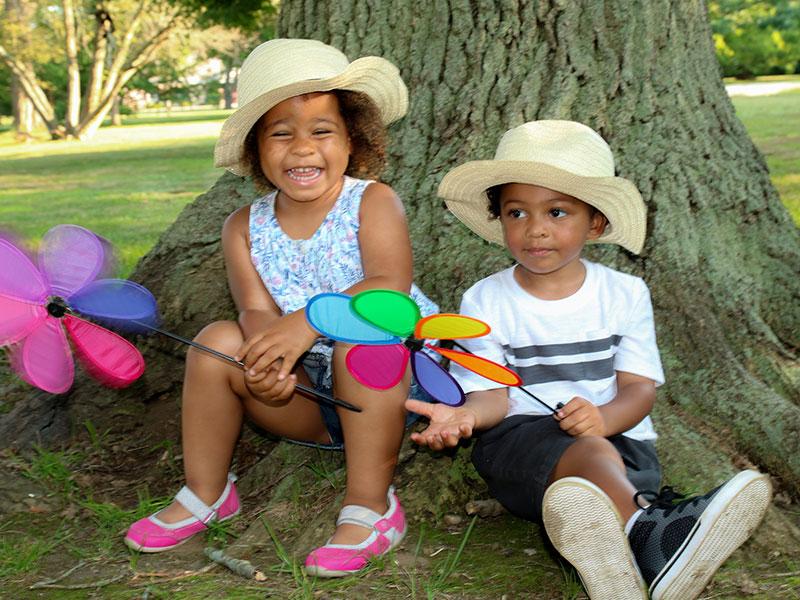 two children sat under a tree with summer hats on and windmills in their hands